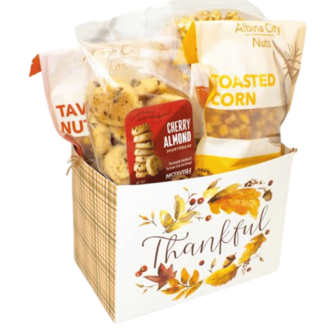 A fall-themed gift basket filled with snacks, including bags of Albina City nuts and toasted corn, a bag of cherry almond cookies, and pumpkin pie caramel corn, set in a white basket with autumn leaves and the word 'Thankful' printed on the front.