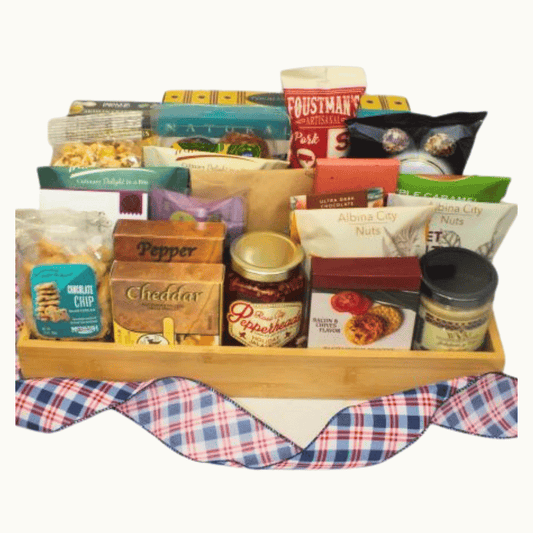 A wooden gift basket filled with assorted snack and food items, including bags of chips, nuts, crackers, and a jar of cheddar cheese, arranged on a white background with a red, white, and blue plaid fabric underneath.
