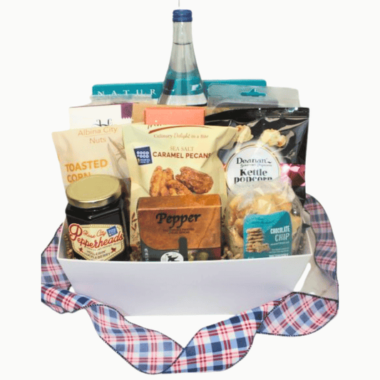 A gourmet gift basket filled with assorted snacks and treats, including a bottle of water, a jar of Pepperheads, toasted corn, caramel pecans, kettle popcorn, and chocolate chip cookies, arranged in a white basket with a red, blue, and white plaid ribbon on a white background.