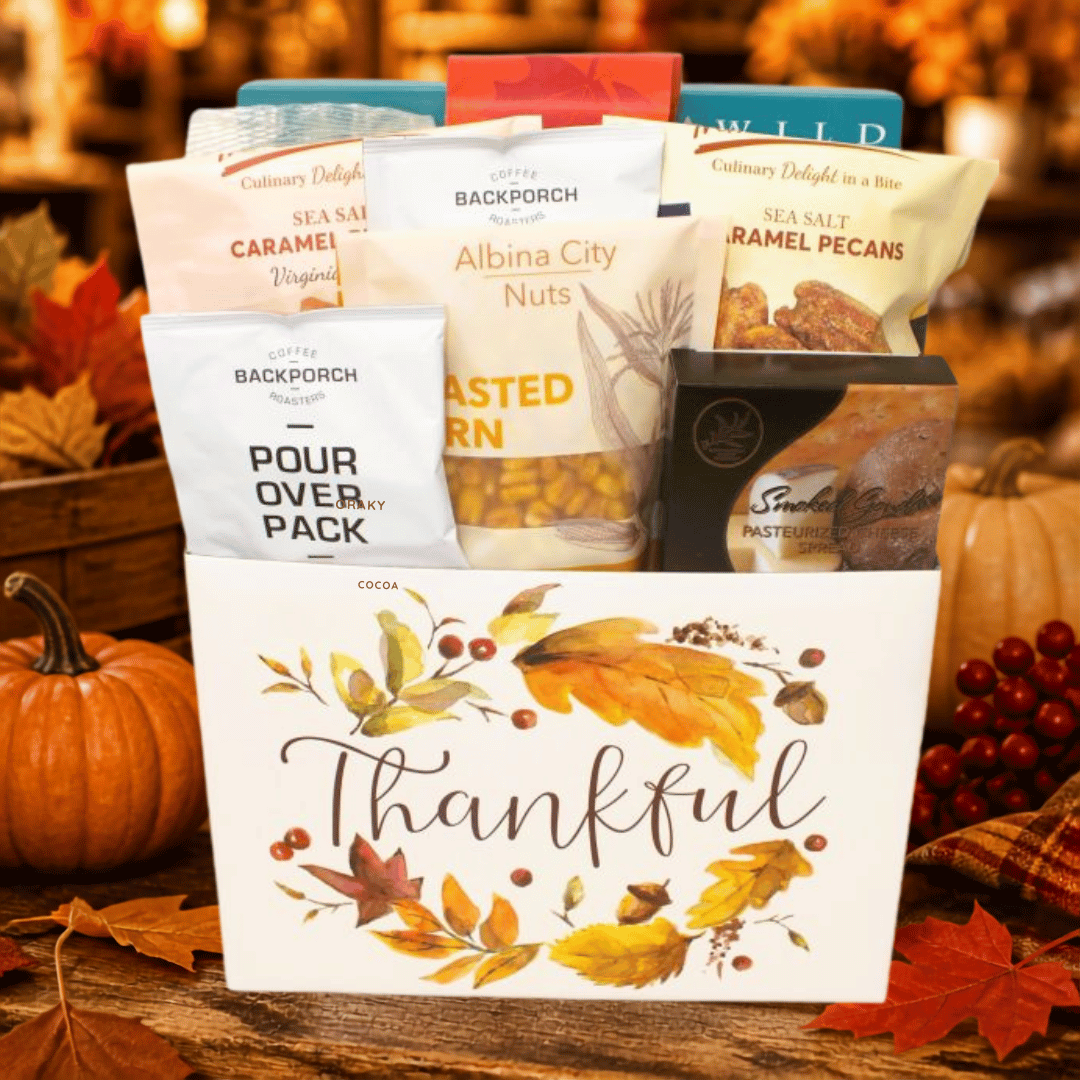 A fall-themed gift basket filled with assorted snack packages, including Backporch Sea Salt Caramel, Albina City Nuts, and Sea Salt Pecans, presented in a white 'Thankful' branded tote basket with autumn leaf illustrations. The basket is styled on a rustic wooden table with pumpkins, red berries, and fall leaves in the background.